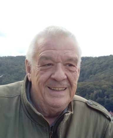 Älterer Mann mit grauen Haaren, lächelnd vor einer leicht bewaldeten Landschaft.