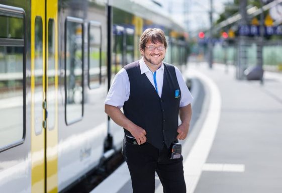 Lächelnder Zugbegleiter in Uniform an einem Bahnhof neben einem Zug.