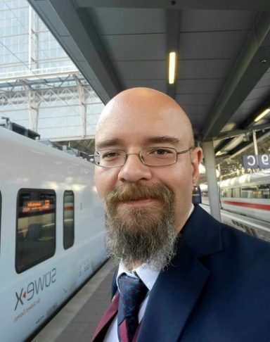 Ein Mann mit Brille und Bart in einem Anzug steht am Bahnsteig neben einem Zug.