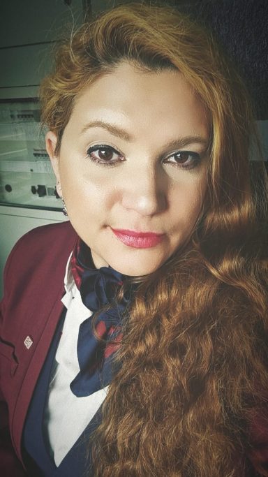 Frau mit langen, lockigen roten Haaren, rotem Lippenstift und blauer Bluse im lässigen Outfit.